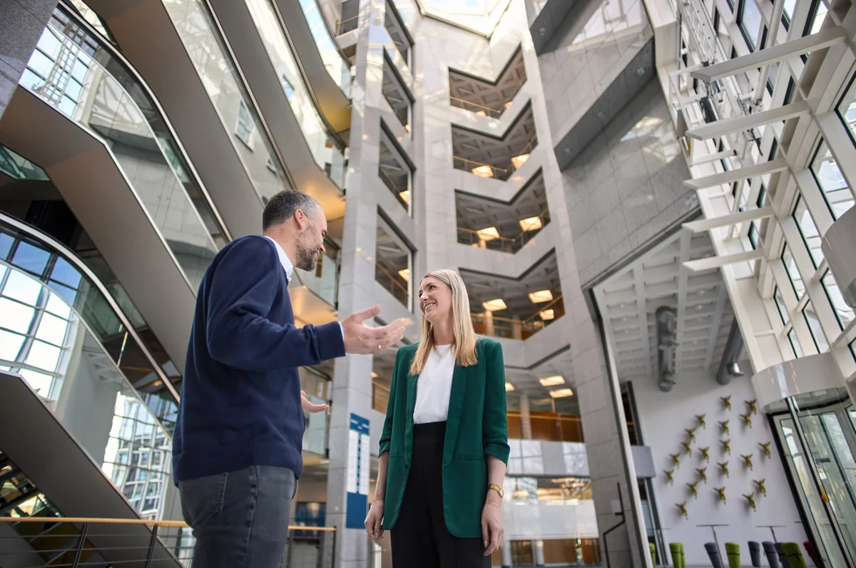 Zwei Personen stehen in einem modernen, hellen Gebäude mit großem Innenraum. Der Mann spricht gestikulierend, während die Frau interessiert zuhört. Beide sind geschäftlich gekleidet und scheinen ein Gespräch über ein wichtiges Thema zu führen.