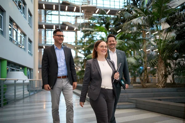 Drei Personen, zwei Männer und eine Frau, gehen in einem modernen Bürogebäude. Sie tragen formelle Kleidung und wirken freundlich. Um sie herum sind große Fenster und Pflanzen, die eine angenehme Atmosphäre schaffen. Die Gruppe sieht aus, als ob sie ein erfolgreiches Gespräch führen.