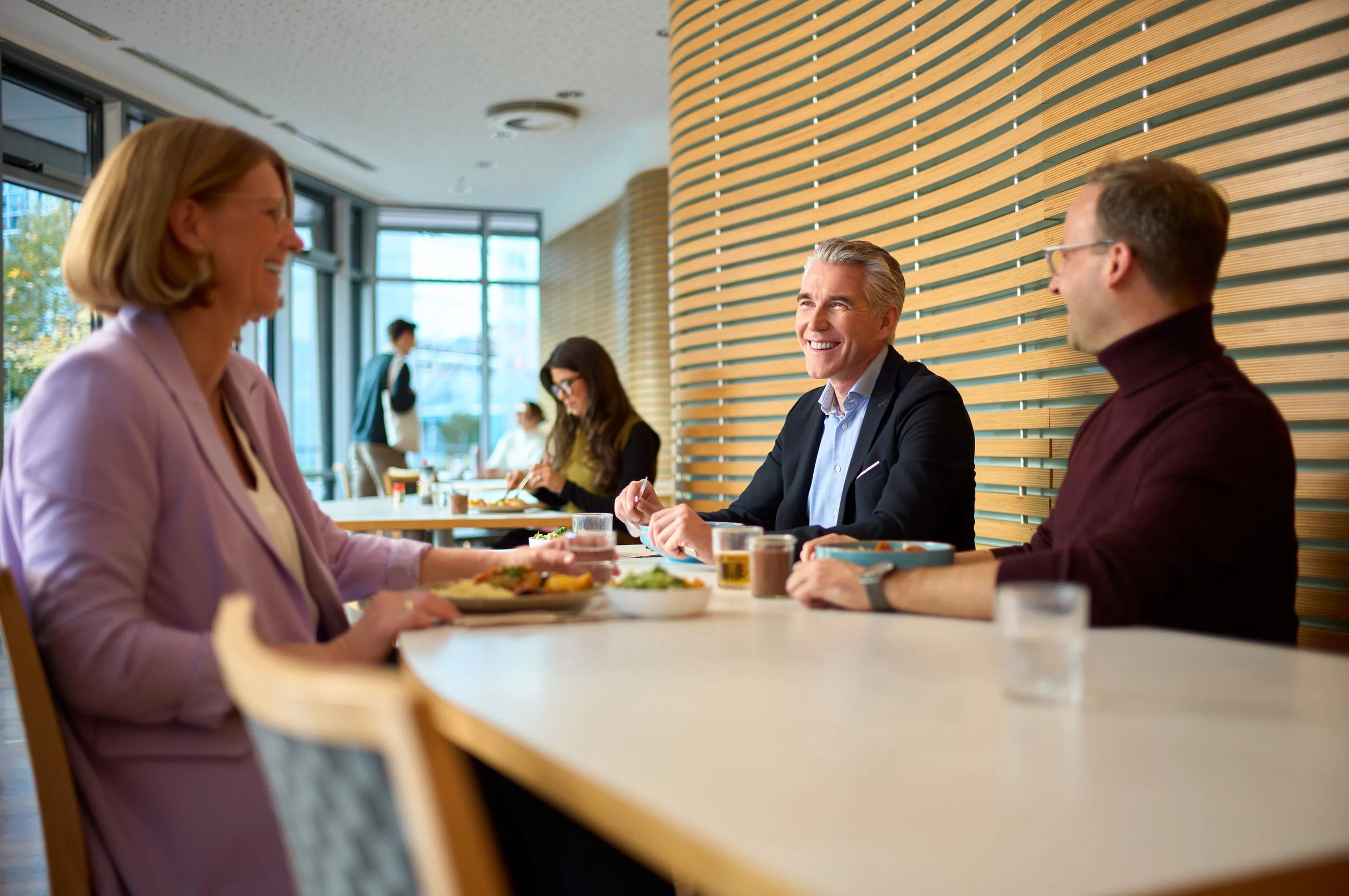 Drei Personen sitzen in einem modernen Café an einem Tisch, unterhalten sich und lachen. Eine Frau links trägt einen lila Blazer, der Mann in der Mitte einen dunklen Anzug, und der Mann rechts einen weinroten Pullover. Sie wirken entspannt und genießen die Atmosphäre.