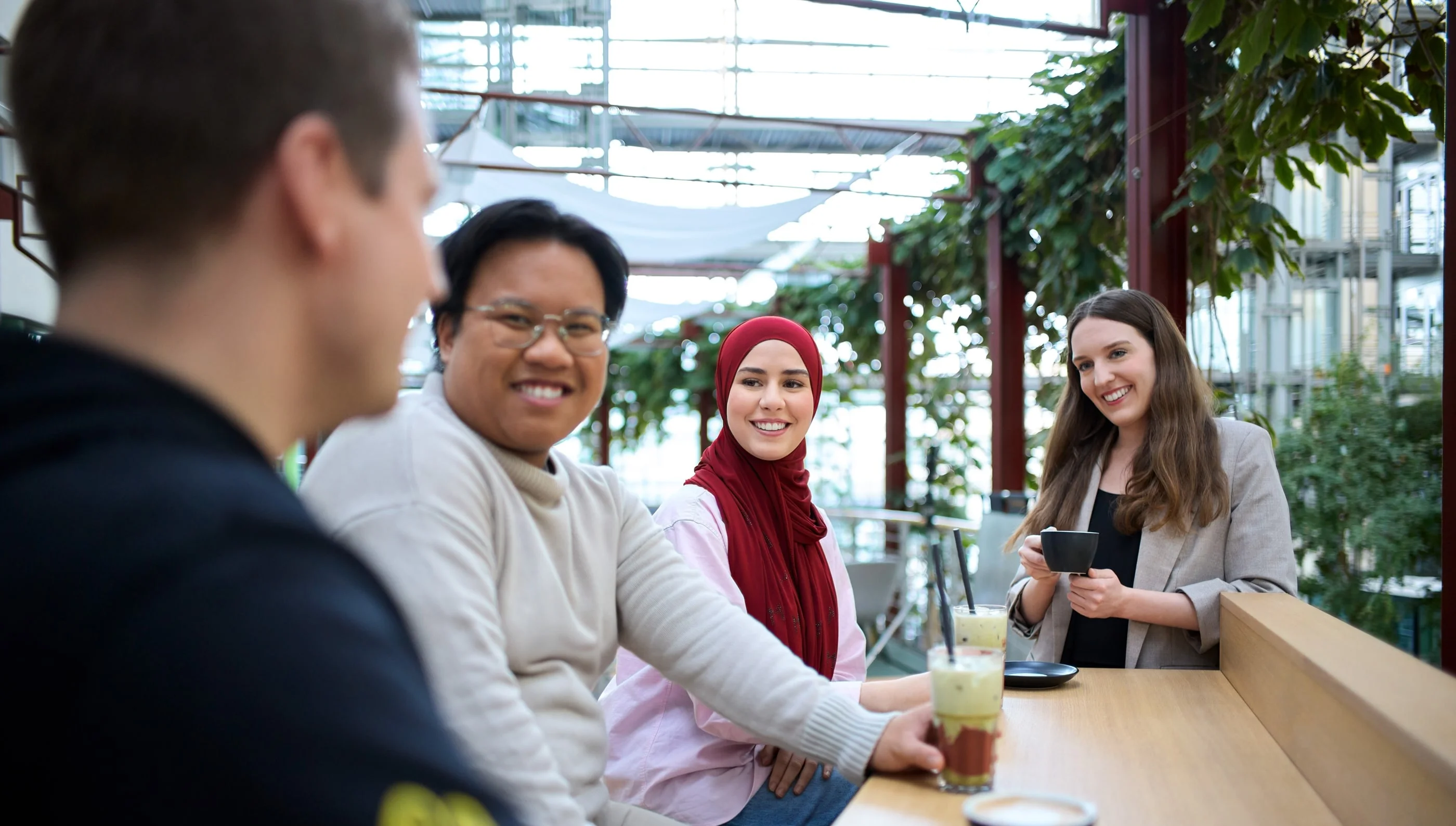 Vier Personen sitzen an einem Tisch in einem hellen, gemütlichen Raum mit vielen Pflanzen. Sie lächeln und sprechen miteinander. Auf dem Tisch stehen verschiedene Getränke, darunter zwei Tassen Kaffee und ein grüner Smoothie.