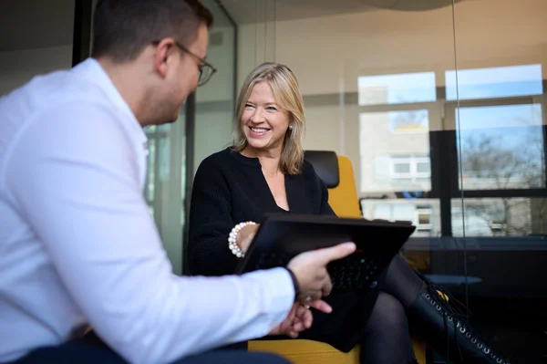 Zwei Personen sitzen in einem modernen Büro. Eine Frau mit blonden Haaren lächelt, während sie auf ein Tablet blickt. Ein Mann im weißen Hemd hält ebenfalls ein Tablet. Die Atmosphäre ist freundlich und einladend.