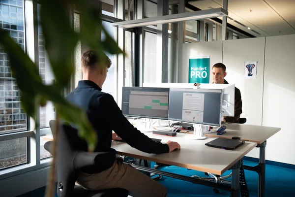 Zwei Personen sitzen an Schreibtischen in einem modernen Büro. Eine Person arbeitet an einem Computer, während die andere ihn anspricht. Im Hintergrund ist ein Schild mit der Aufschrift "Hundert PRO" zu sehen. Es gibt große Fenster und eine Pflanze im Vordergrund.