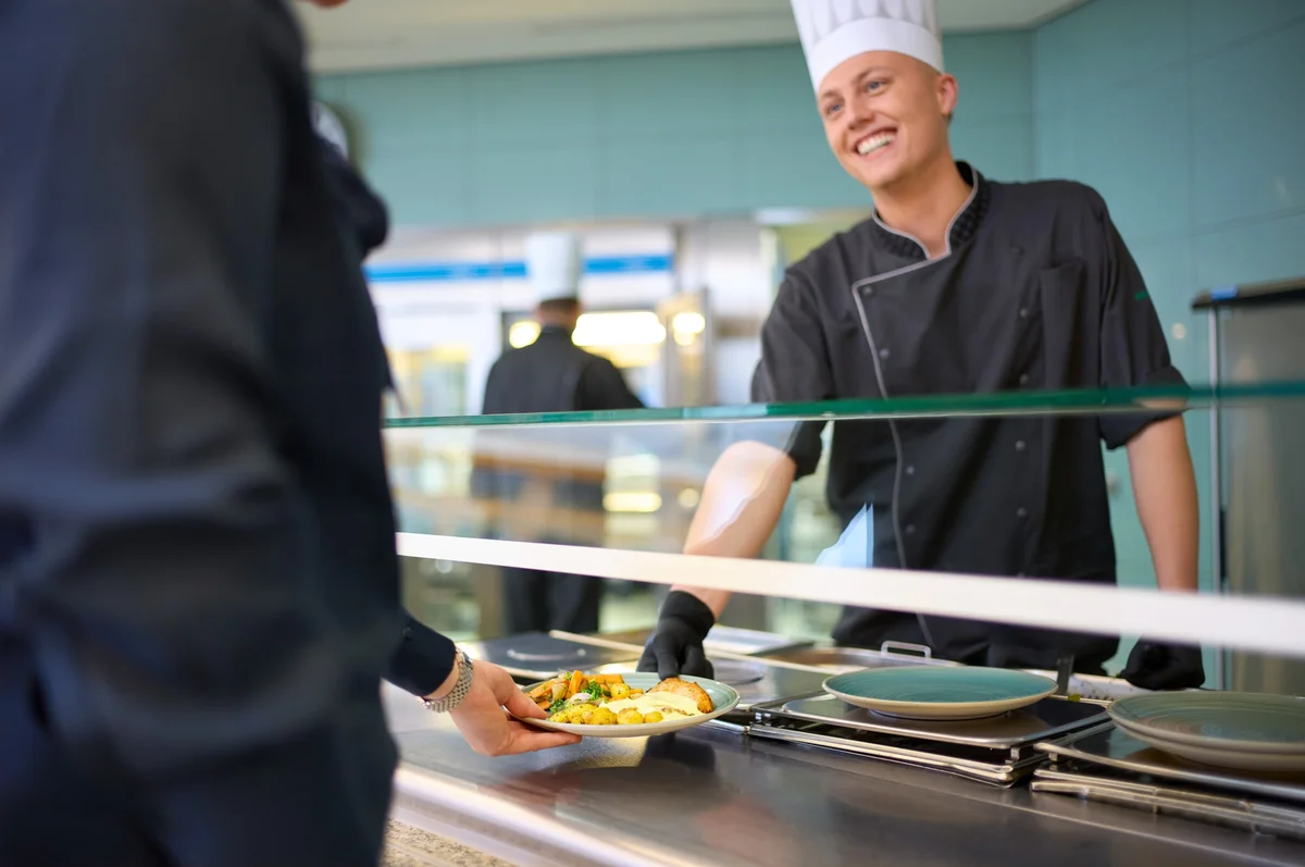 Ein lächelnder Koch in einer Schulkantine reicht einem Gast einen Teller mit Essen. Der Koch trägt eine Schürze und eine Kochmütze, während der Gast mit einer Hand den Teller entgegennimmt. Im Hintergrund sind zusätzliche Geschirr und eine Glasfront sichtbar.