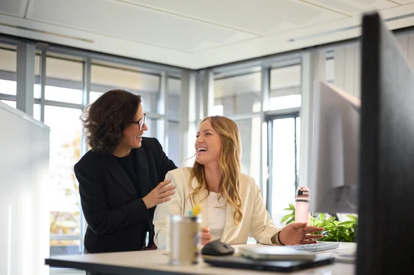 Zwei Frauen in einem modernen Büro sitzen an einem Schreibtisch. Eine Frau lächelt und gestikuliert, während die andere sie unterstützend an der Schulter berührt. Im Hintergrund sind Pflanzen und ein Computer zu sehen, was eine freundliche Arbeitsumgebung vermittelt.