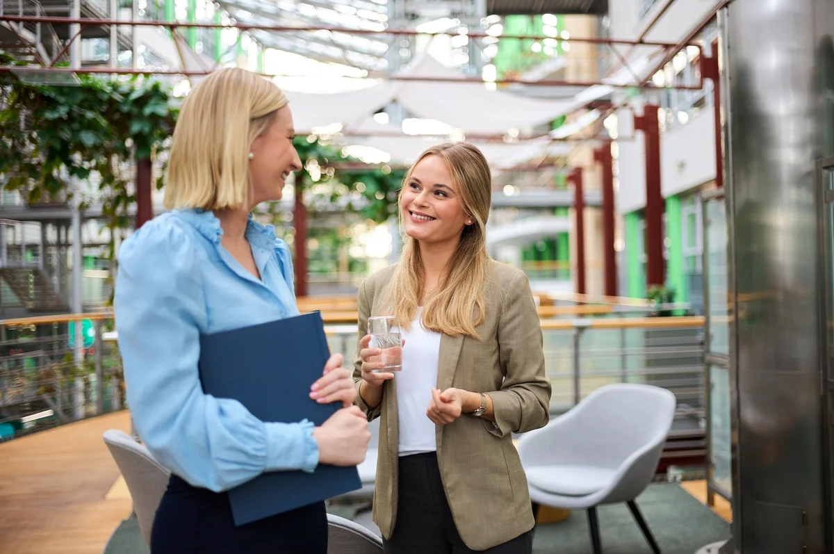 Zwei Frauen stehen in einem modernen, hellen Innenraum und unterhalten sich. Eine Frau hält einen blauen Ordner, die andere hat ein Glas Wasser in der Hand. Beide lächeln und wirken freundlich, während sie miteinander sprechen.