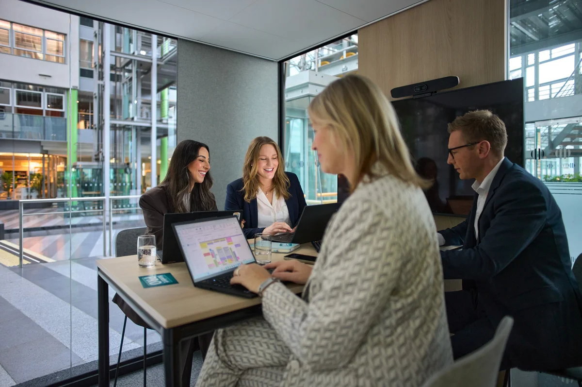 Eine Gruppe von vier Personen sitzt an einem Tisch in einem modernen Büro. Sie arbeiten an Laptops und diskutieren. Die Atmosphäre ist freundlich und produktiv, mit viel natürlichem Licht und einer zeitgemäßen Umgebung.