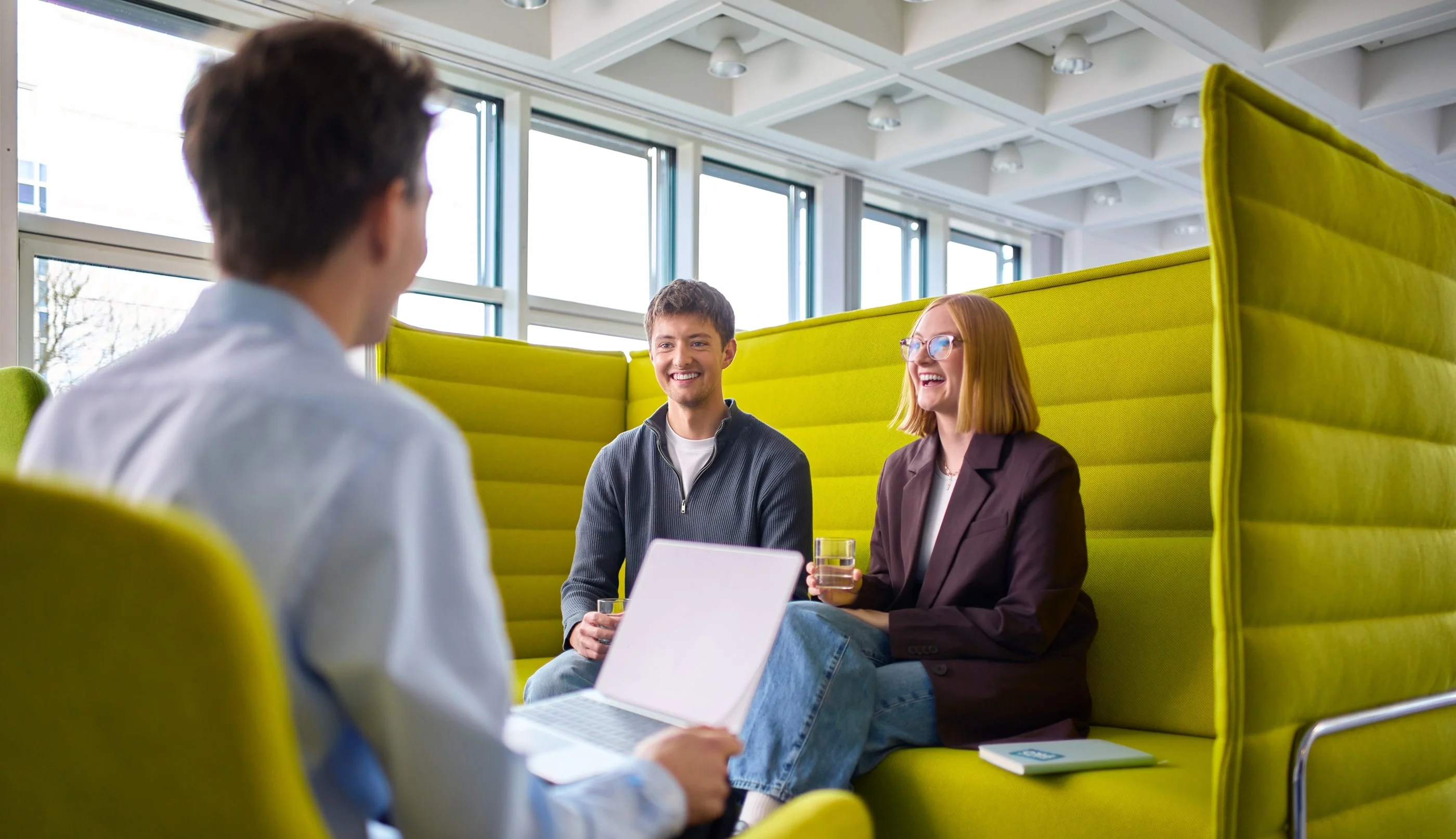 Drei Personen sitzen in einem hellen, modernen Raum, umgeben von grünen Sitzgelegenheiten. Zwei der Personen, ein Mann und eine Frau, lächeln und scheinen an einem Gespräch teilzunehmen, während die dritte Person, die ihnen gegenüber sitzt, Notizen macht.