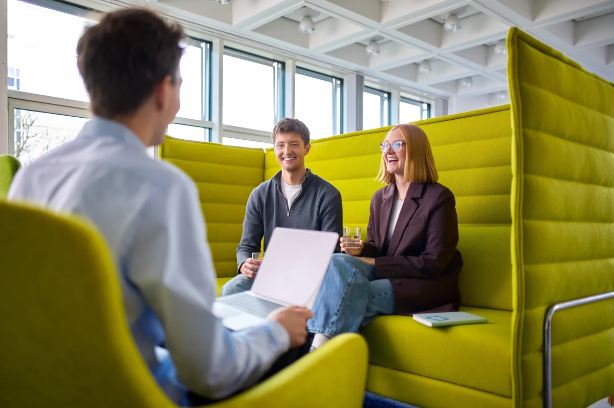 Drei Personen sitzen in einem hellen, modernen Raum, umgeben von grünen Sitzgelegenheiten. Zwei der Personen, ein Mann und eine Frau, lächeln und scheinen an einem Gespräch teilzunehmen, während die dritte Person, die ihnen gegenüber sitzt, Notizen macht.