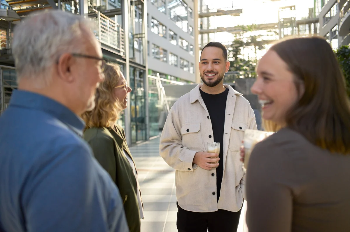 Zwei Frauen und zwei Männer stehen in einem modernen Innenraum und unterhalten sich. Alle wirken freundlich und entspannt. Jeder hält ein Getränk in der Hand, während sie lachen und sich austauschen. Sonnenlicht fällt durch große Fenster und schafft eine angenehme Atmosphäre.