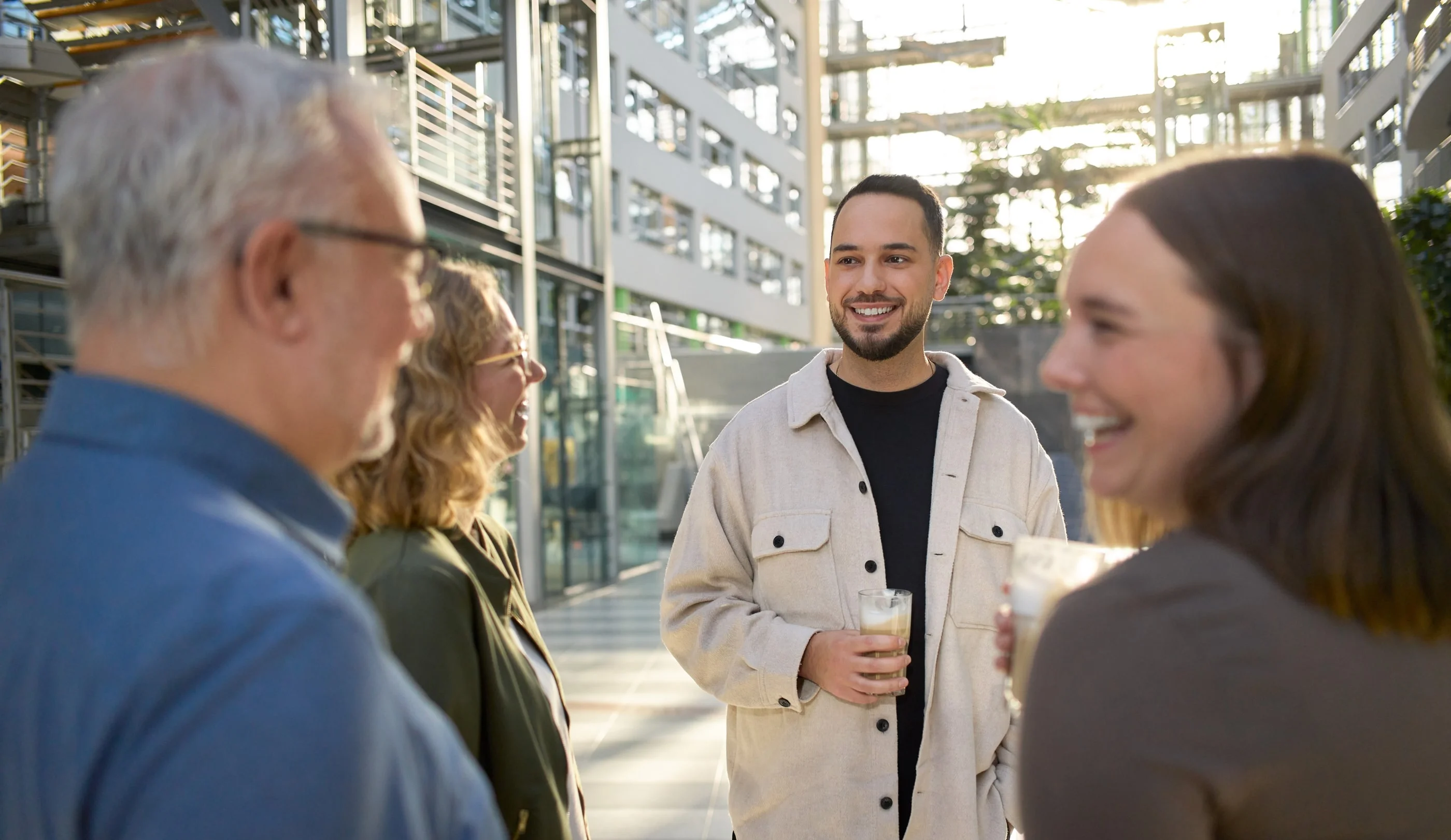 Zwei Frauen und zwei Männer stehen in einem modernen Innenraum und unterhalten sich. Alle wirken freundlich und entspannt. Jeder hält ein Getränk in der Hand, während sie lachen und sich austauschen. Sonnenlicht fällt durch große Fenster und schafft eine angenehme Atmosphäre.