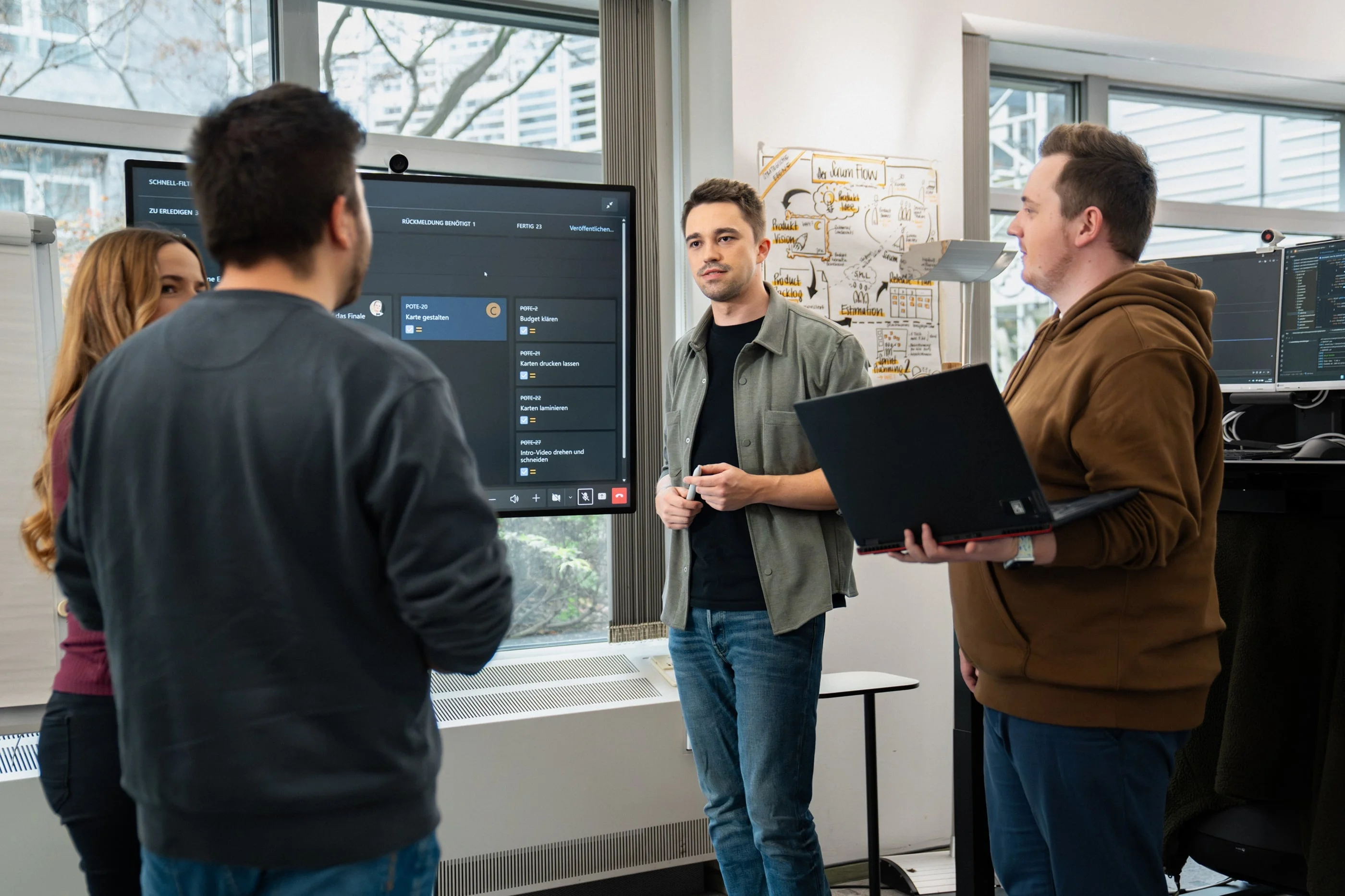 Eine Gruppe von vier Personen steht um einen Bildschirm in einem modernen Büro. Eine Person erklärt etwas, während die anderen aufmerksam zuhören. Ein Mann hält einen Laptop in der Hand.