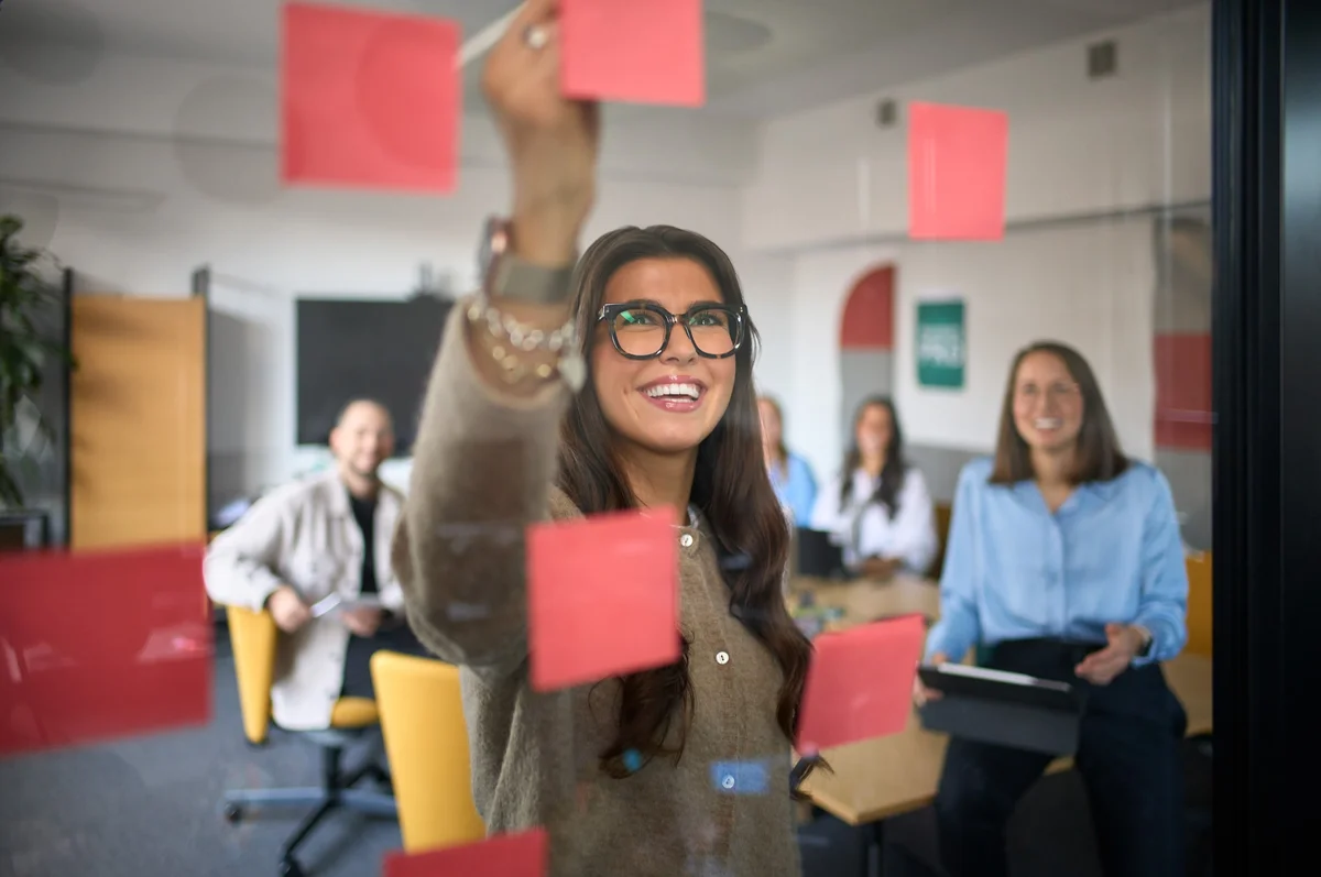 Eine lächelnde Frau mit Brille befestigt Haftnotizen an einer Glaswand. Im Hintergrund sitzen vier Personen, die ihr aufmerksam zuschauen, während sie an einem kreativen Meeting teilnehmen. Die Atmosphäre wirkt konzentriert und inspirierend.