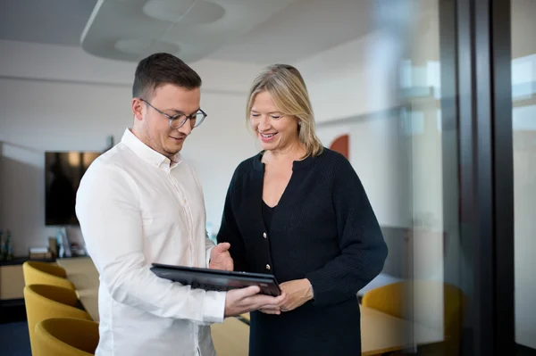 Zwei Personen stehen in einem modernen Büroraum und betrachten gemeinsam ein Tablet. Der Mann trägt ein weißes Hemd und Brille, während die Frau in einem schwarzen Oberteil lächelt. Die Atmosphäre wirkt freundlich und kollegial.