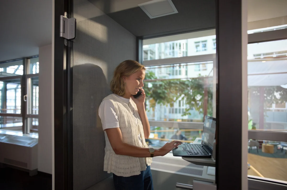 Eine Frau steht in einem Telefonkabine und spricht am Telefon. Sie hat ihren Laptop auf einer Ablage und tippt gleichzeitig. Im Hintergrund sind Fenster mit natürlichem Licht und grüne Pflanzen sichtbar. Die Szene vermittelt eine ruhige, produktive Arbeitsatmosphäre.
