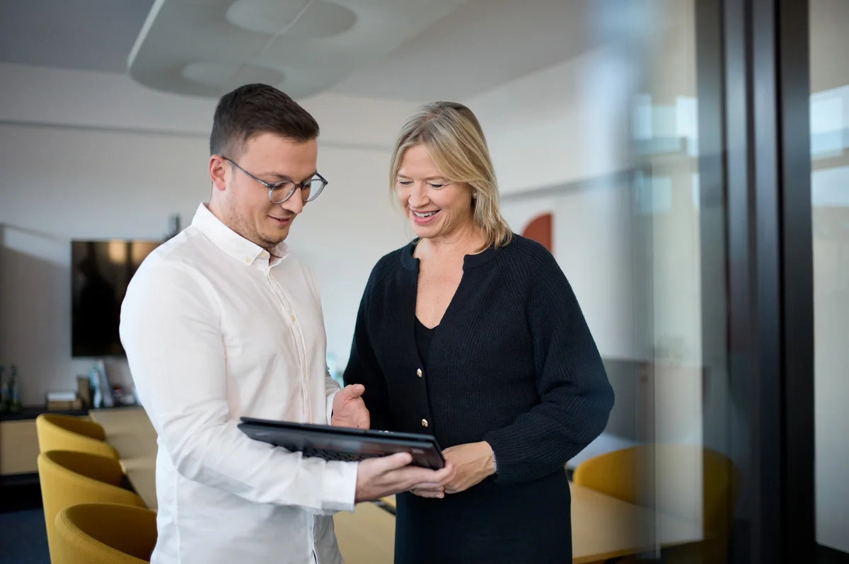 Ein junger Mann und eine Frau stehen in einem modernen Büro und betrachten gemeinsam ein Tablet. Der Mann trägt eine Brille und ein weißes Hemd, die Frau hat blonde Haare und trägt eine schwarze Bluse. Beide wirken freundlich und engagiert.