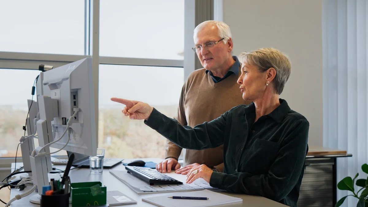 Ein älterer Mann und eine ältere Frau stehen an einem Schreibtisch und schauen auf einen Computerbildschirm. Die Frau zeigt mit dem Finger auf den Bildschirm, während der Mann interessiert zusieht. Beide scheinen ein Gespräch über den Inhalt des Bildschirms zu führen.