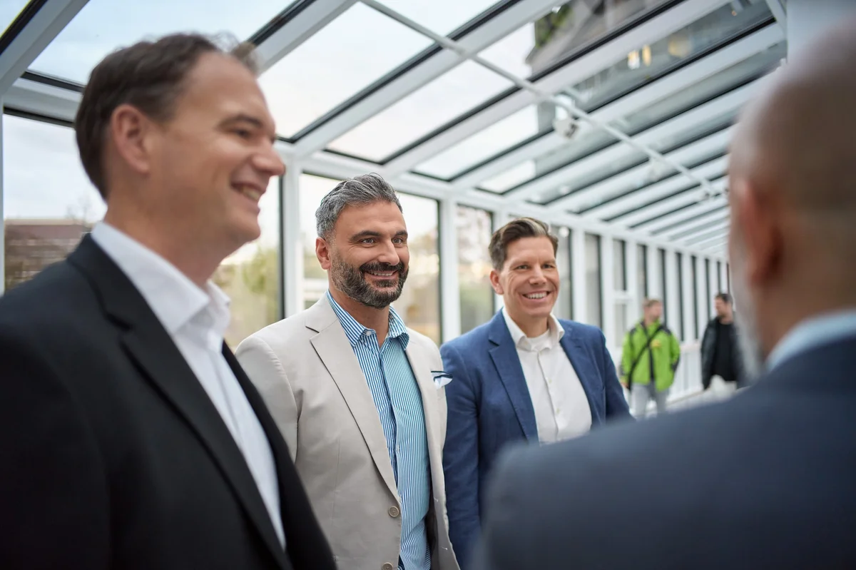 Vier Männer stehen in einem hellen, modernen Raum mit vielen Fenstern. Sie lächeln und scheinen ein Gespräch zu führen. Der Mann links trägt einen schwarzen Anzug, der in der Mitte einen hellen Blazer, und der Mann rechts einen blauen Anzug.