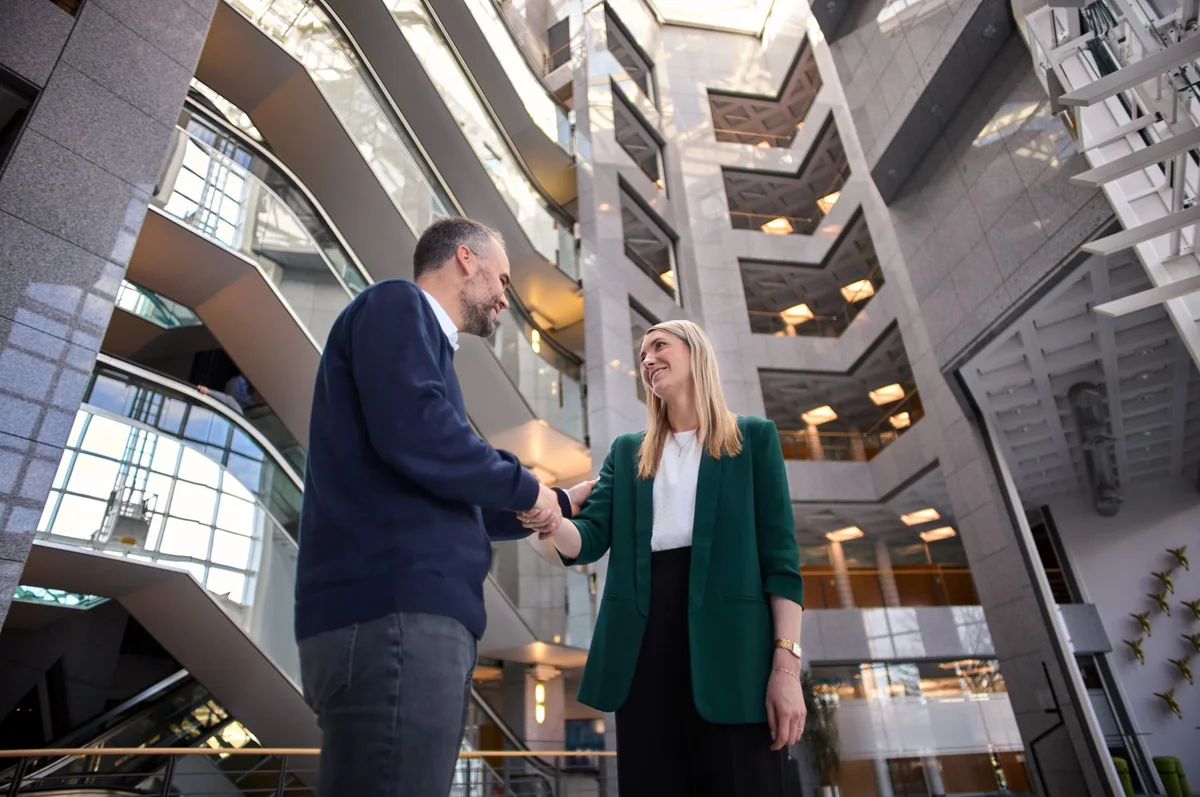 Zwei Geschäftspersonen schütteln sich die Hände in einem modernen Bürogebäude. Der Mann trägt einen dunkelblauen Pullover und die Frau eine grüne Jacke. Sie stehen in einem offenen Raum mit Treppen und einem hellen, einladenden Ambiente.