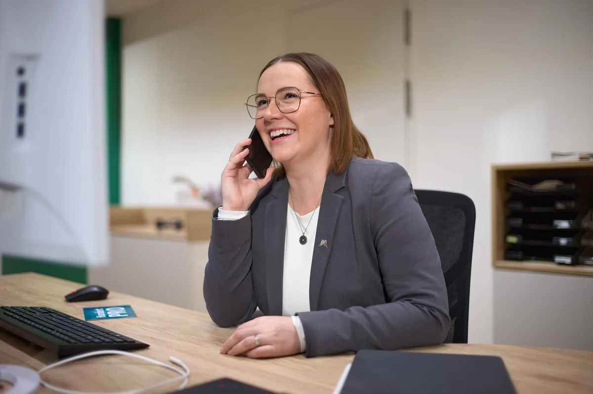 Eine fröhliche Frau sitzt an einem Schreibtisch, spricht am Telefon und lächelt. Sie trägt eine Brille und einen grauen Blazer. Auf dem Tisch sind ein Computer und einige Unterlagen sichtbar. Der Hintergrund ist hell und modern.