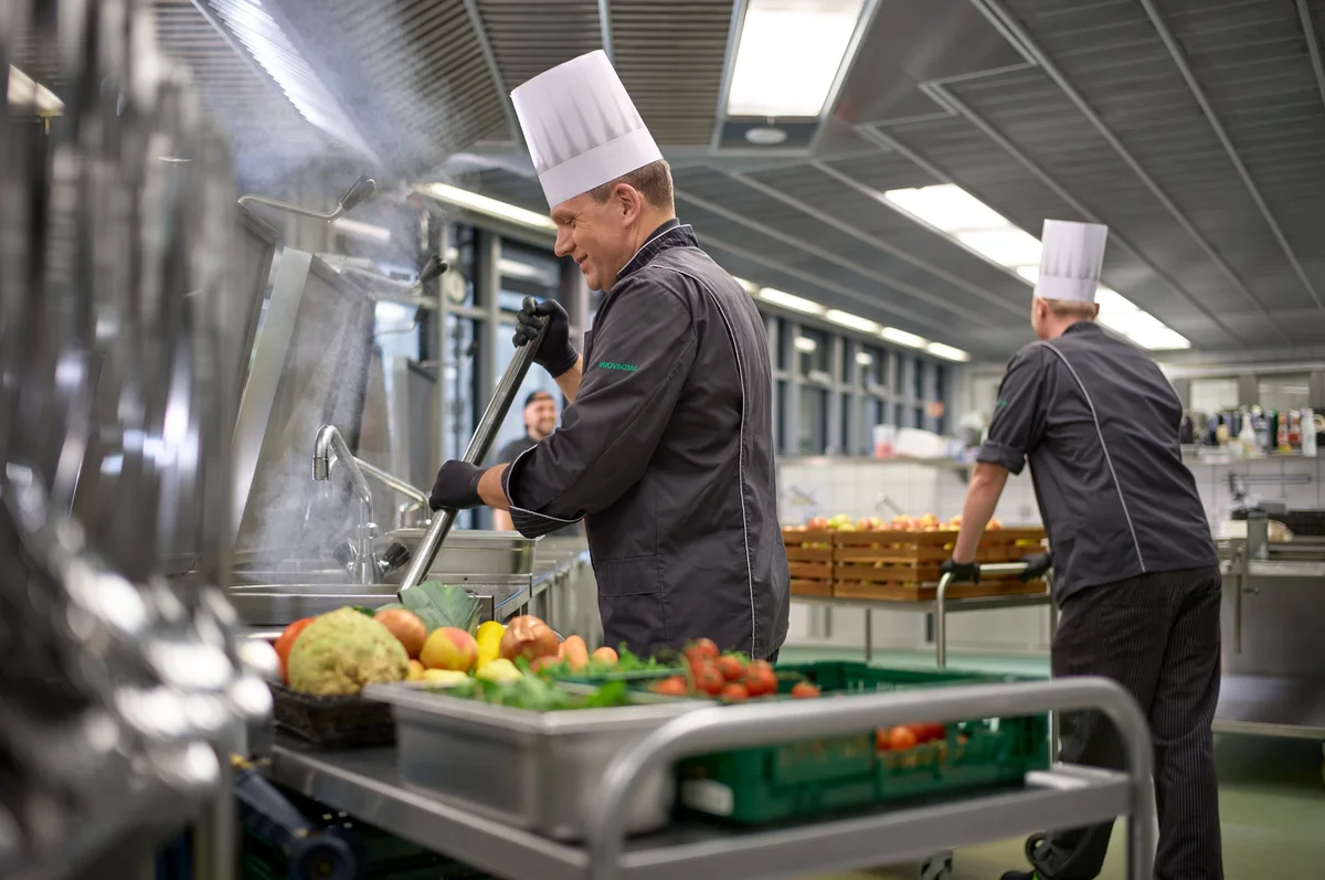 Zwei Köche in professioneller Küche sind mit der Zubereitung von Speisen beschäftigt. Einer rührt in einem großen Topf, während der andere im Hintergrund steht. Vor ihnen liegt eine Auswahl frischer Zutaten wie Gemüse und Obst auf einem Wagen.