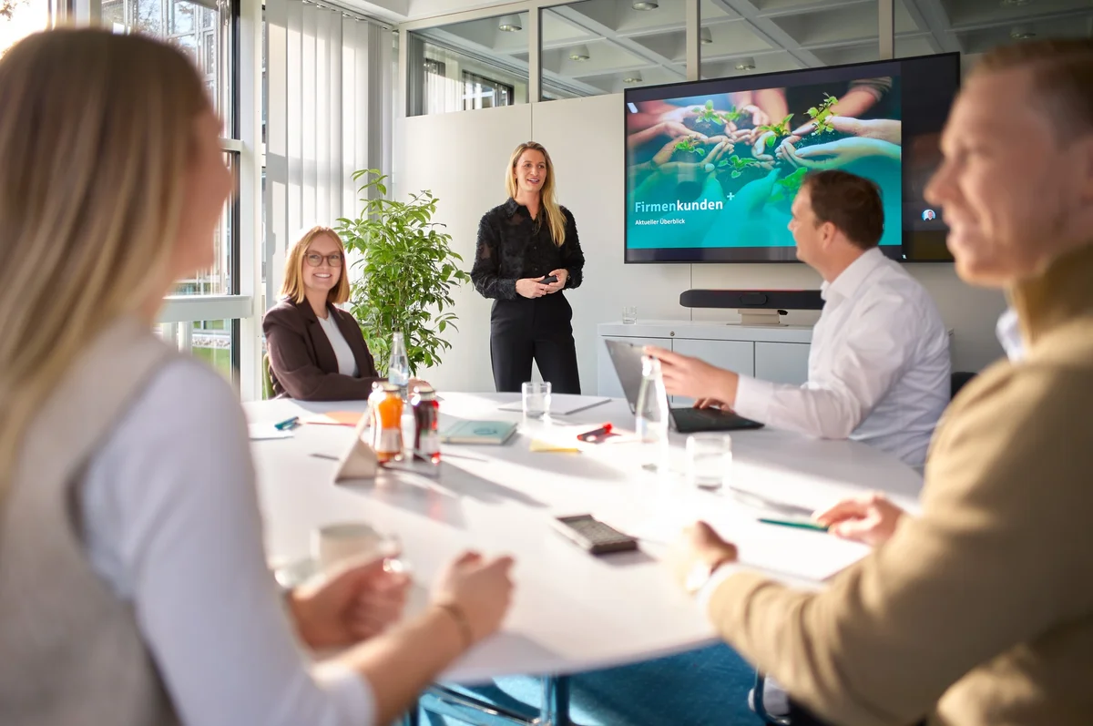 Eine Präsentation in einem Konferenzraum. Eine Frau steht vorne und spricht, während fünf Personen am Tisch aufmerksam zuhören. Im Hintergrund ist ein Bildschirm mit einer Präsentation über "Firmenkunden" sichtbar. Es gibt Getränke und Notizen auf dem Tisch.