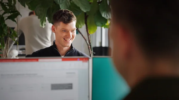 Ein junger Mann mit einem freundlichen Lächeln sitzt hinter einem Computerbildschirm. Im Hintergrund ist eine Pflanze sichtbar und eine andere Person scheint an einem Arbeitsplatz zu sein. Der Mann wirkt zufrieden und engagiert in einer Gesprächssituation.