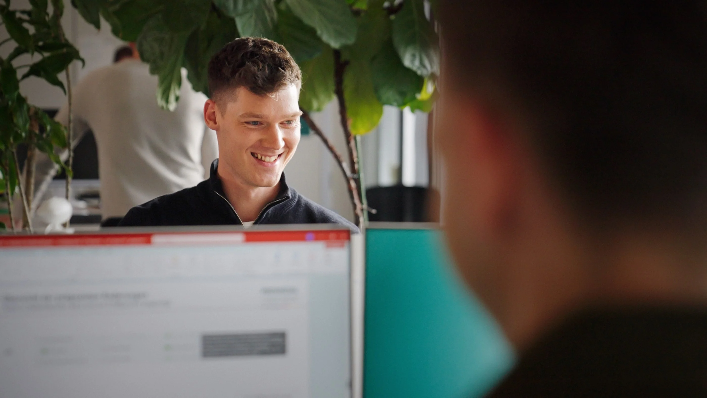 Ein junger Mann mit einem freundlichen Lächeln sitzt hinter einem Computerbildschirm. Im Hintergrund ist eine Pflanze sichtbar und eine andere Person scheint an einem Arbeitsplatz zu sein. Der Mann wirkt zufrieden und engagiert in einer Gesprächssituation.