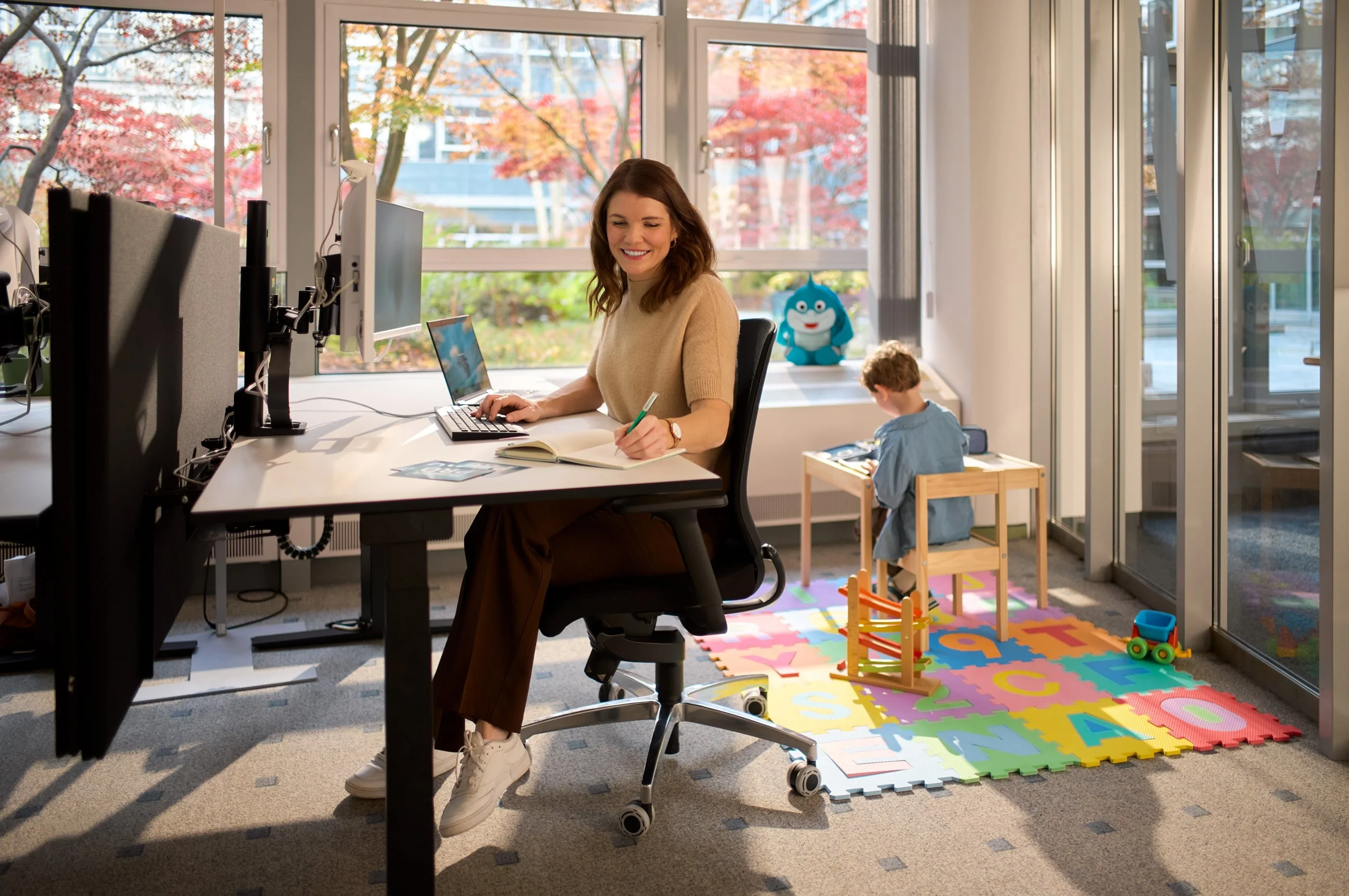 Eine Frau sitzt lächelnd an einem Schreibtisch mit Laptop und Notizbuch. Hinter ihr steht ein kleiner Junge an einem Tisch, der mit Spielzeug beschäftigt ist. Große Fenster im Hintergrund lassen viel Tageslicht herein, und draußen sind Bäume mit buntem Laub zu sehen.