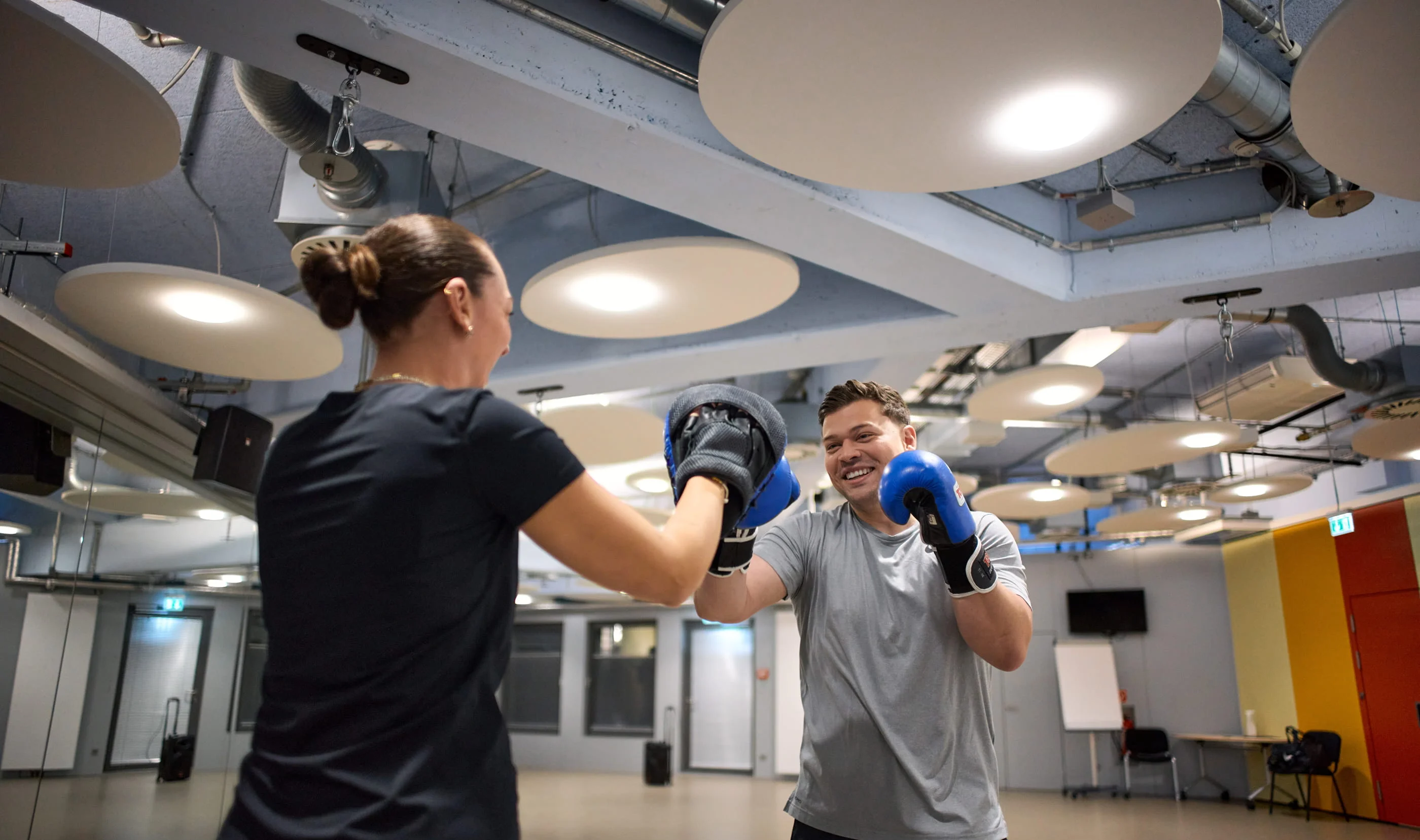 Ein Mann in grauem Shirt und blauen Boxhandschuhen trainiert mit einer Frau in schwarzem Shirt in einem Fitnessraum. Sie tragen beide Boxhandschuhe und lächeln, während sie üben.