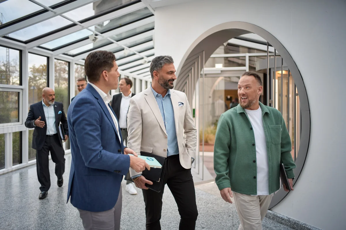 Drei Männer unterhalten sich lachend in einem modernen Gang mit Glasdecken. Im Hintergrund sind weitere Personen sichtbar, die dem sich untereinander unterhalten.
