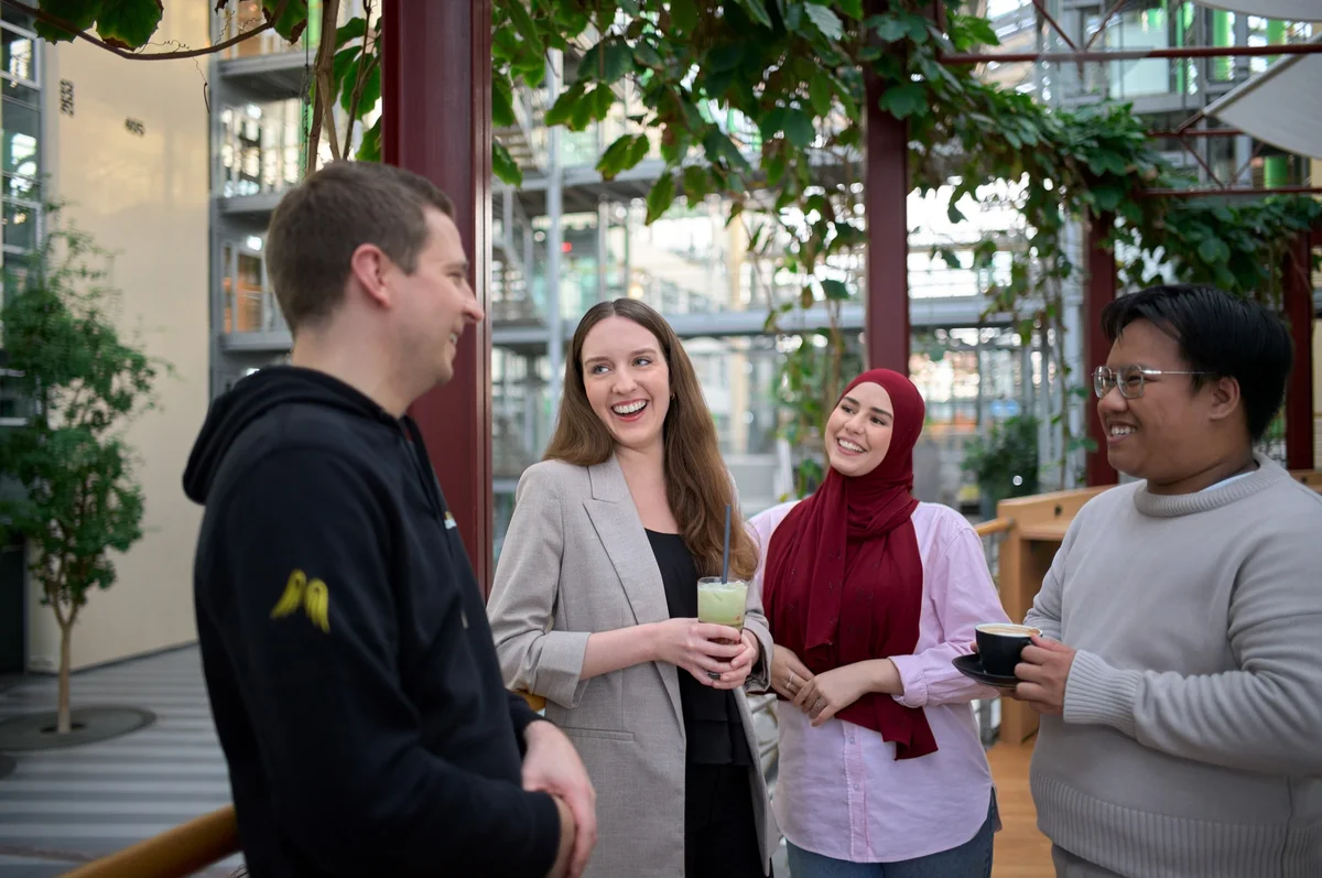 Eine Gruppe von vier Personen steht in einem modernen Innenraum mit Pflanzen. Sie lachen und unterhalten sich, während eine Frau ein Getränk hält. Ihre Kleidung ist lässig und vielfältig, was eine lockere, freundliche Atmosphäre vermittelt.