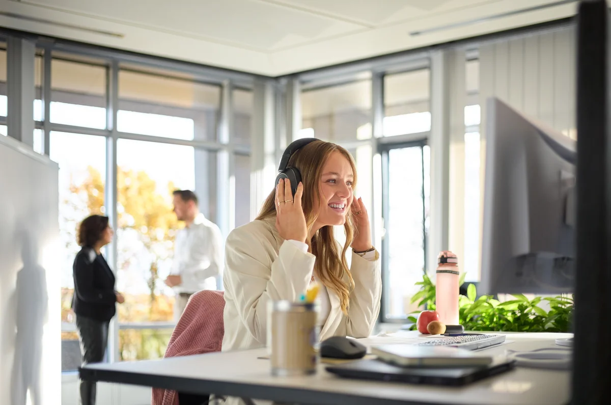 Eine junge Frau sitzt an einem Schreibtisch in einem modernen Büro, trägt Kopfhörer und lächelt. Sie scheint an einem Gespräch oder einer Videokonferenz teilzunehmen. Im Hintergrund sind zwei Personen zu sehen, die miteinander sprechen. Pflanzen und Getränke sind auf dem Tisch sichtbar.