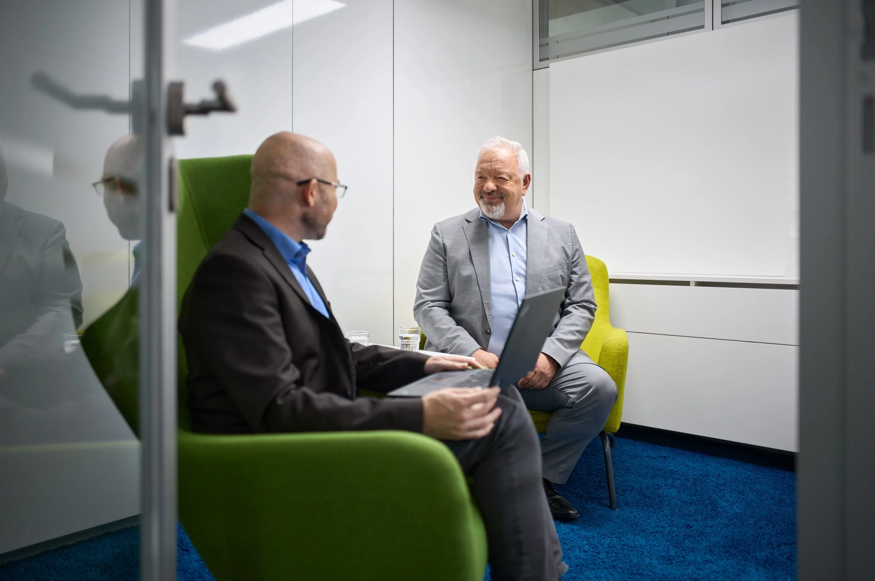 Zwei Männer sitzen in einem modernen Büro. Beide sitzen in einem grünen Stuhl und einer hält einen Laptop, während der andere lächelt. Beide wirken in einem freundlichen Austausch vertieft.