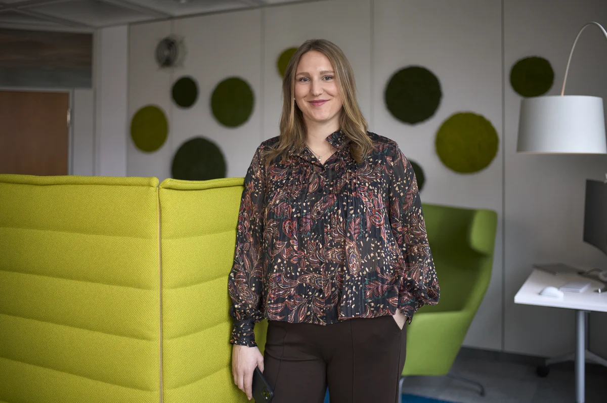 Eine Frau steht lächelnd in einem modernen Büro mit grünen Akustik-Paneelen an der Wand. Sie trägt eine gemusterte Bluse und hält ein Handy in der Hand. Im Hintergrund sind Schreibtische und ein grüner Sessel zu sehen.