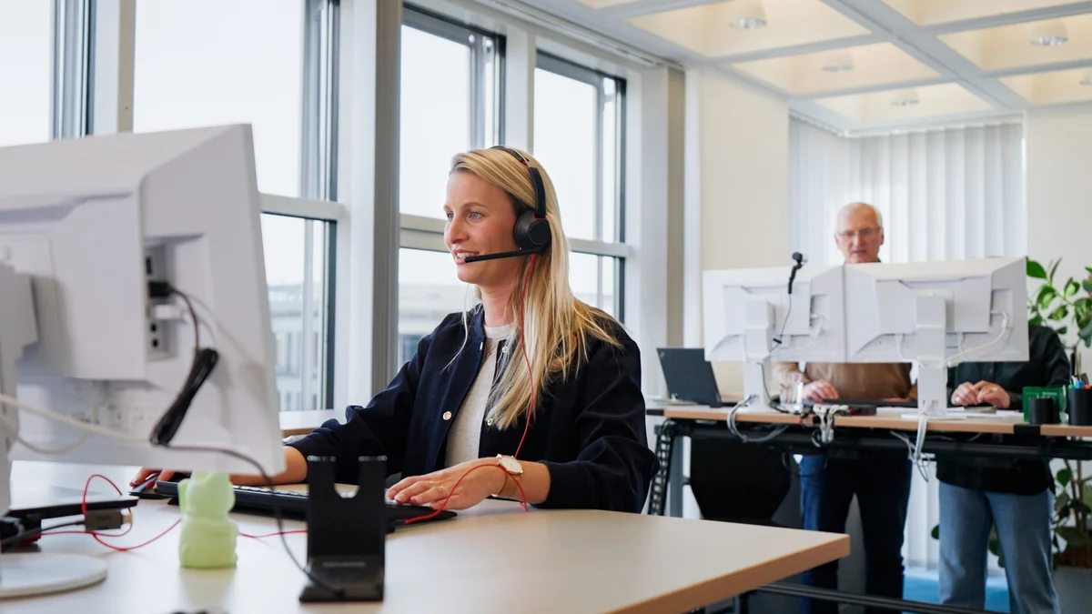 Eine blonde Frau trägt ein Headset und arbeitet an einem Computer. Im Hintergrund stehen zwei Personen an weiteren Arbeitsplätzen, ebenfalls am Computer. Der Raum ist hell und modern gestaltet, mit großen Fenstern und Pflanzen.