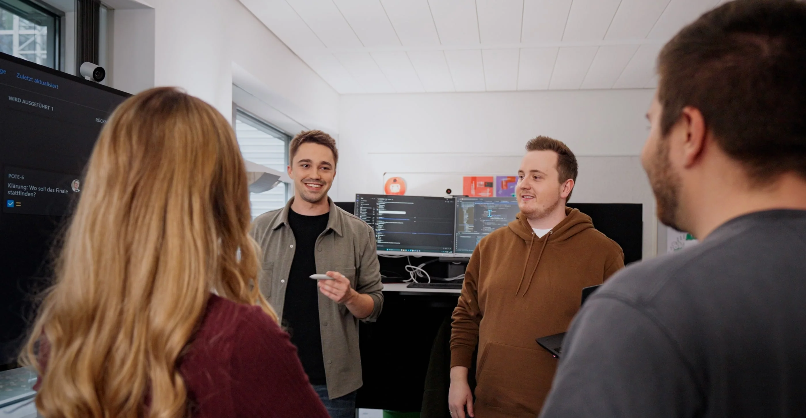 Vier Personen in einem modernen Büroraum unterhalten sich. Ein Mann steht im Mittelpunkt und spricht, während die anderen aufmerksam zuhören. Auf einem Monitor im Hintergrund sind Computer-Codes sichtbar, was auf ein technisches Thema hindeutet.