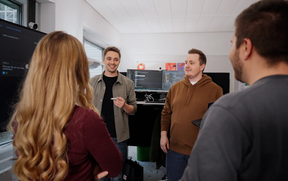 Vier Personen in einem modernen Büroraum unterhalten sich. Ein Mann steht im Mittelpunkt und spricht, während die anderen aufmerksam zuhören. Auf einem Monitor im Hintergrund sind Computer-Codes sichtbar, was auf ein technisches Thema hindeutet.