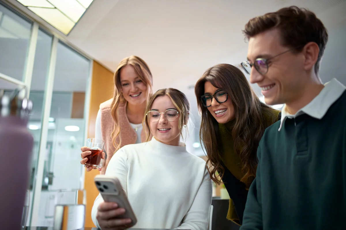 Vier junge Erwachsene stehen zusammen und schauen auf ein Smartphone. Eine Frau in der Mitte lächelt, während die anderen gespannt auf den Bildschirm blicken. Die Atmosphäre ist freundlich und entspannt.