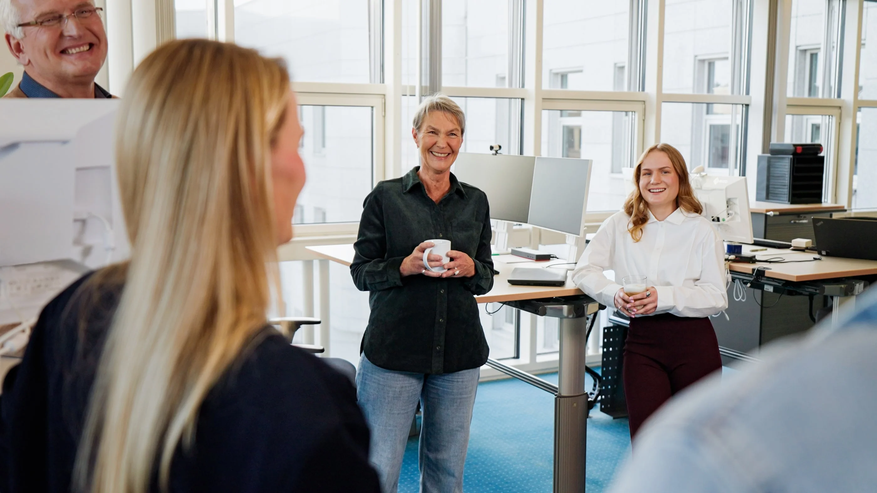 Eine Gruppe von drei Personen im Büro steht zusammen und lächelt. Zwei Frauen halten Kaffeetassen, während die dritte Person, ein Mann, im Hintergrund steht und ebenfalls lächelt. Die Atmosphäre ist freundlich und entspannt.