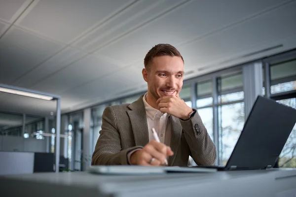 Ein junger Mann sitzt an einem Schreibtisch in einem modernen Büro und schaut lächelnd auf seinen Laptop. Er hat einen Stift in der Hand und wirkt nachdenklich, während er eine Idee notiert oder an einer Aufgabe arbeitet.