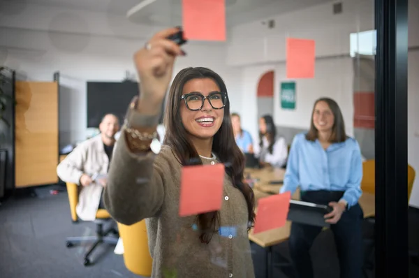 Eine Teilnehmerin in einem Meeting befestigt einen Zettel an einer Glaswand. Im Hintergrund sitzen weitere Personen, die aufmerksam zuschauen. Die Atmosphäre wirkt kollaborativ und engagiert.