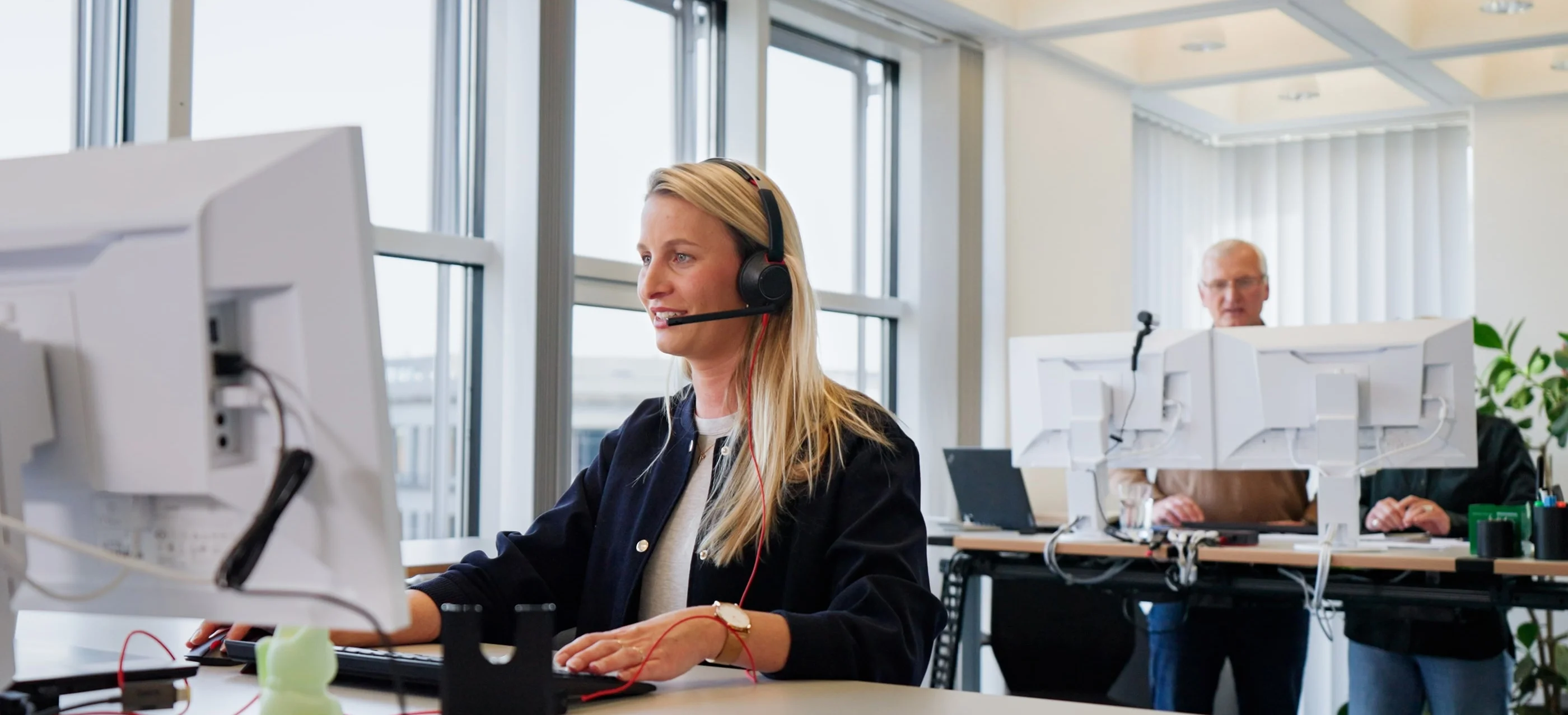 Eine Frau mit Kopfhörern sitzt vor einem Computerbildschirm und arbeitet konzentriert. Im Hintergrund stehen zwei Personen an aufrechten Arbeitsplätzen mit weiteren Computern. Das Büro wirkt modern und hell, mit viel Tageslicht.