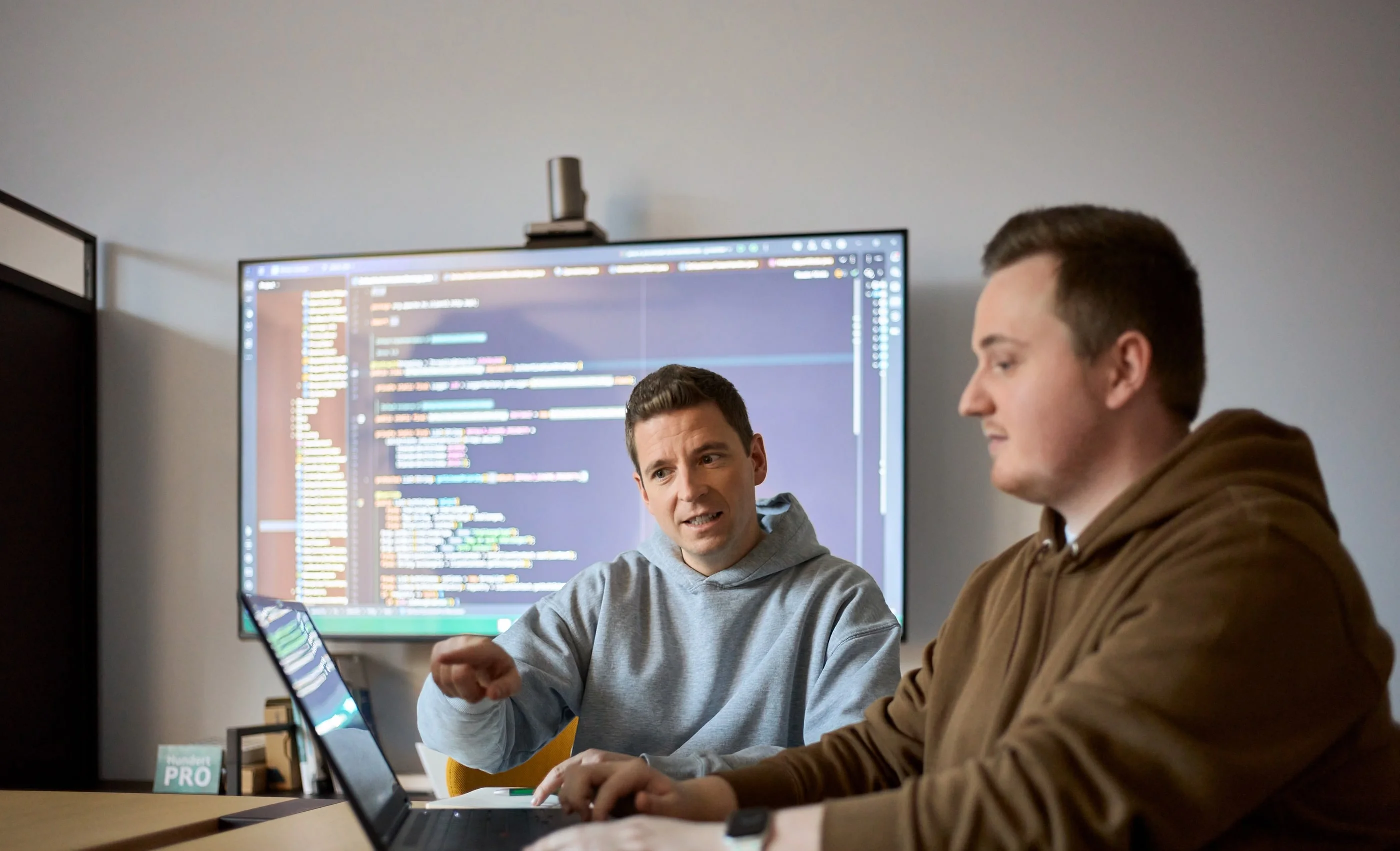 Zwei Männer sitzen an einem Tisch, einer spricht und zeigt auf einen Laptop, während der andere aufmerksam zuhört. Im Hintergrund ist ein Bildschirm mit Programmiercode sichtbar. Die Szene vermittelt einen Austausch über technische Themen oder Softwareentwicklung.