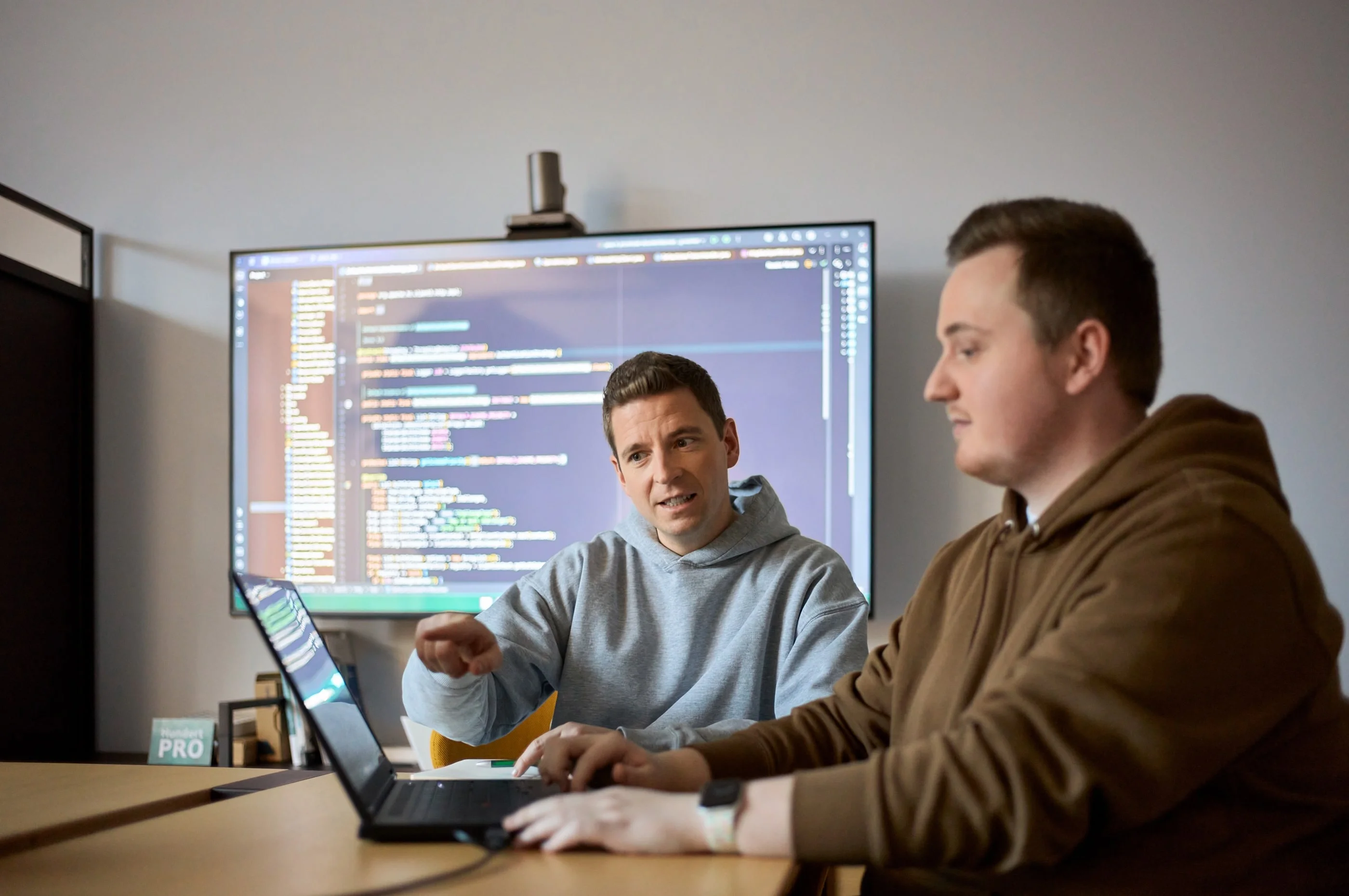 Zwei Männer sitzen an einem Tisch, einer spricht und zeigt auf einen Laptop, während der andere aufmerksam zuhört. Im Hintergrund ist ein Bildschirm mit Programmiercode sichtbar. Die Szene vermittelt einen Austausch über technische Themen oder Softwareentwicklung.