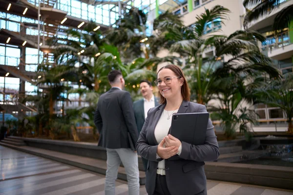 Eine selbstbewusste Frau mit Brille und Tablet steht in einem modernen Innenraum mit Pflanzen. Im Hintergrund sind zwei Männer zu sehen, die miteinander sprechen. Die gesamte Atmosphäre wirkt geschäftlich und freundlich.