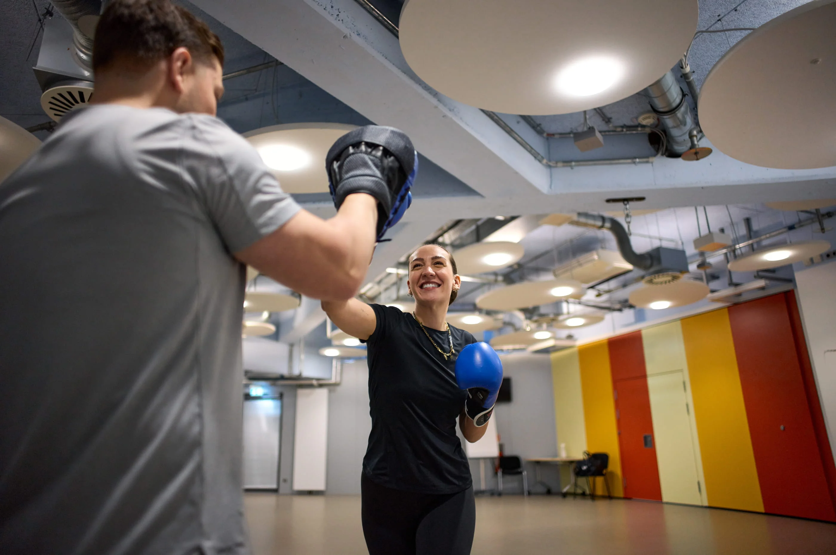 In einem Fitnessstudio trainiert eine Frau mit blauen Boxhandschuhen mit einem Mann, der Pratzen trägt. Beide lachen.