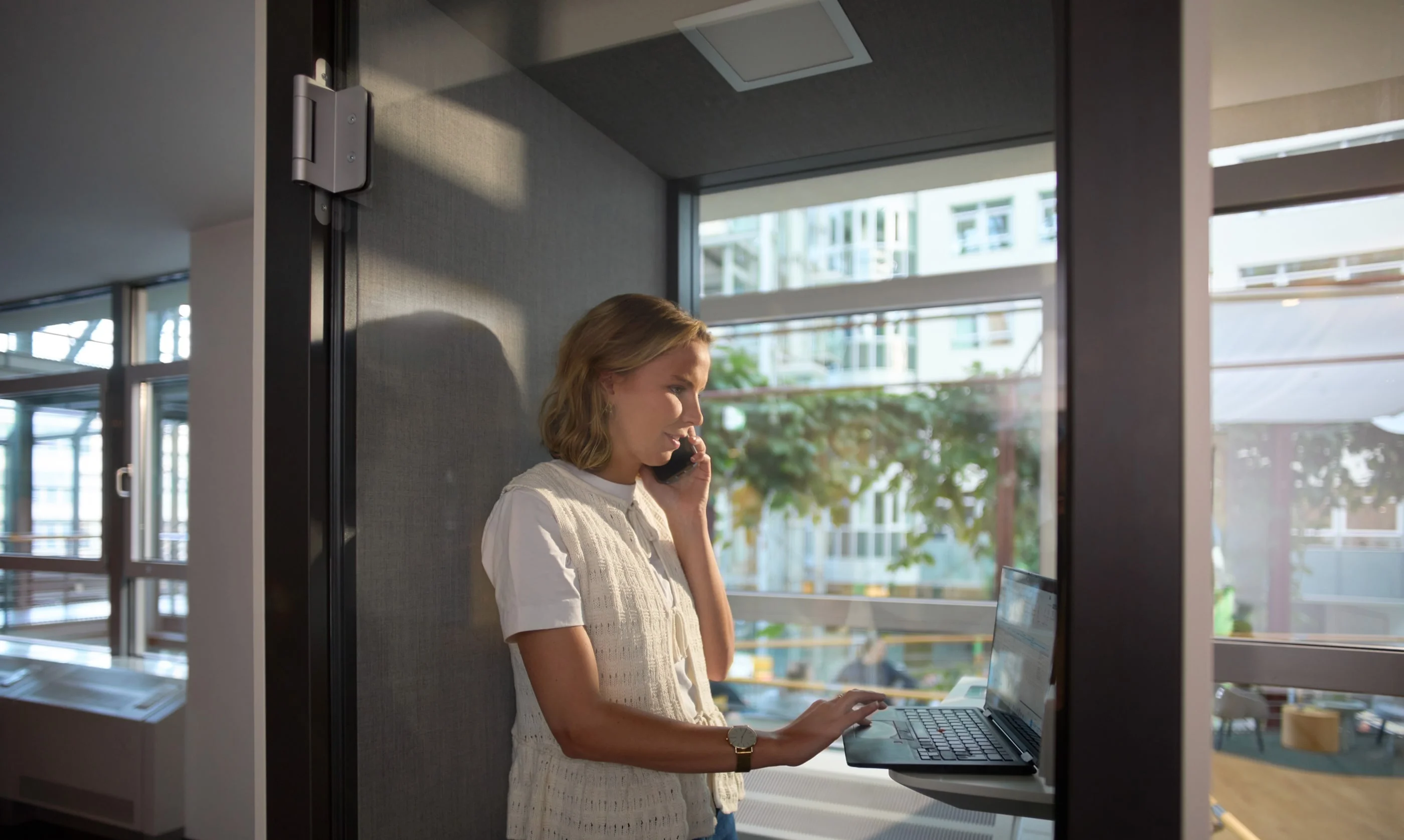 Eine Frau steht in einem kleinen Büro und telefoniert, während sie an einem Laptop arbeitet. Sie trägt ein weißes Oberteil und eine lockere Frisur. Durch die Fenster ist Tageslicht sichtbar, das den Raum erhellt.