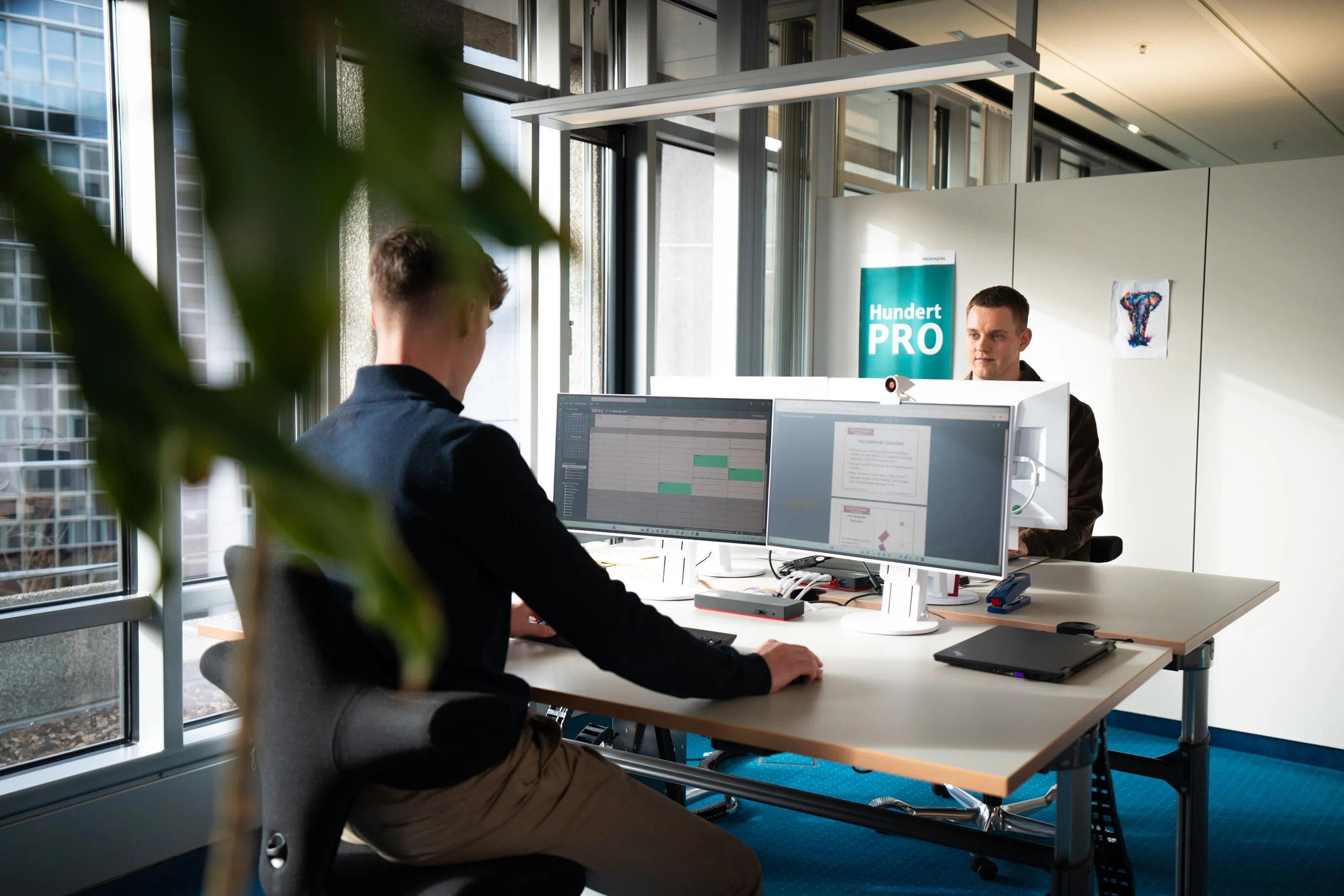 Zwei Personen sitzen an Schreibtischen in einem modernen Büro. Der vordere Mann arbeitet an einem Computer, während der andere auf ihn schaut. Im Hintergrund ist ein Schild mit der Aufschrift "Hundert PRO" zu sehen. Tageslicht strahlt durch große Fenster.