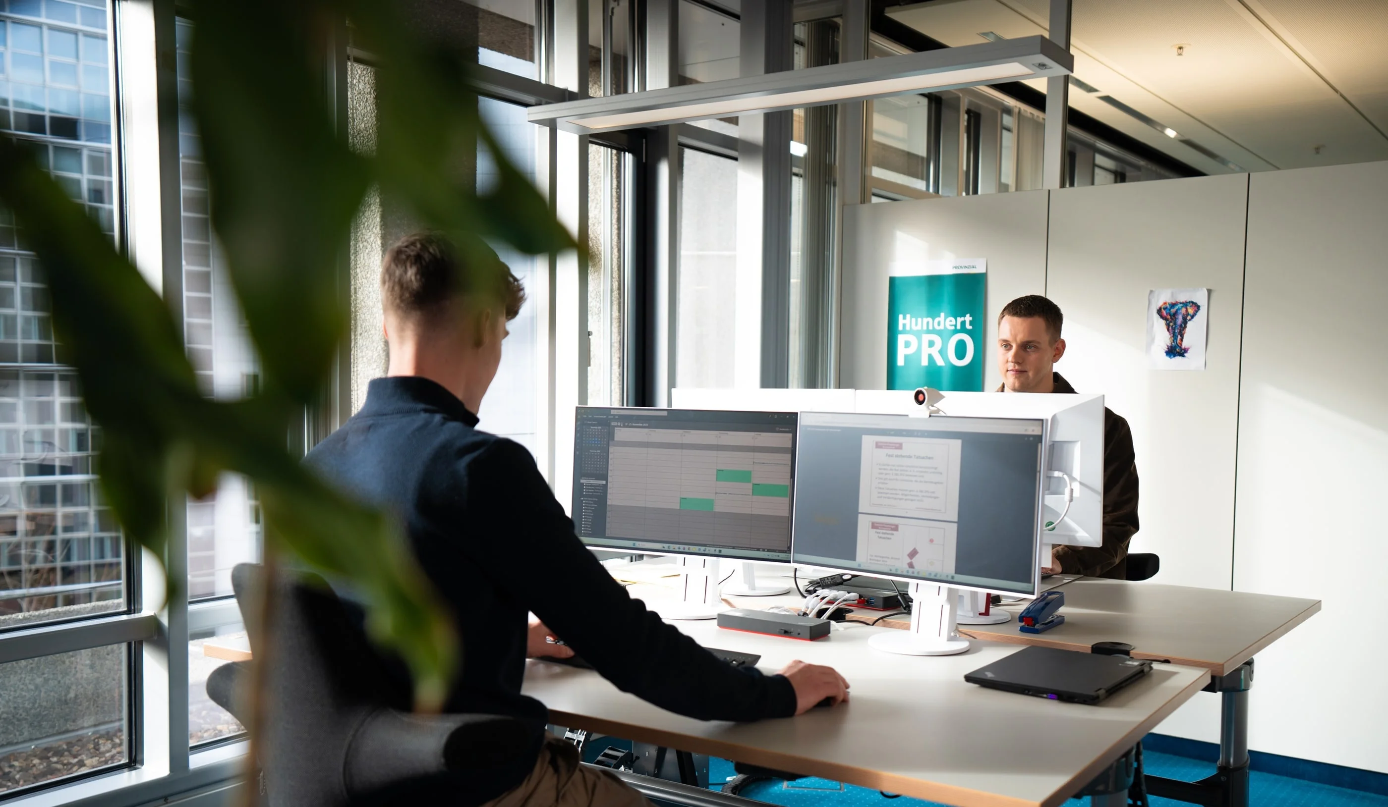Zwei Personen sitzen an Schreibtischen in einem modernen Büro. Der vordere Mann arbeitet an einem Computer, während der andere auf ihn schaut. Im Hintergrund ist ein Schild mit der Aufschrift "Hundert PRO" zu sehen. Tageslicht strahlt durch große Fenster.