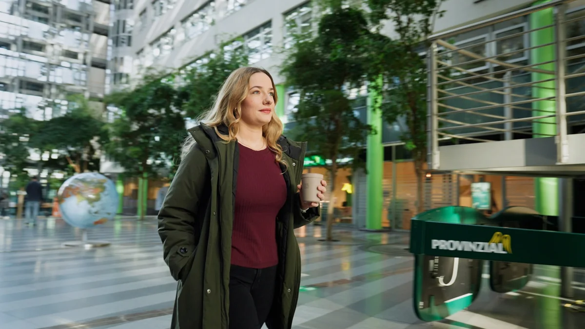 Eine blonde Frau in einem grünen Mantel geht durch ein modernes Gebäude mit großen Fenstern und Pflanzen. Sie hält einen Kaffeebecher in der Hand und schaut entspannt. Im Hintergrund ist eine Weltkugel zu sehen.